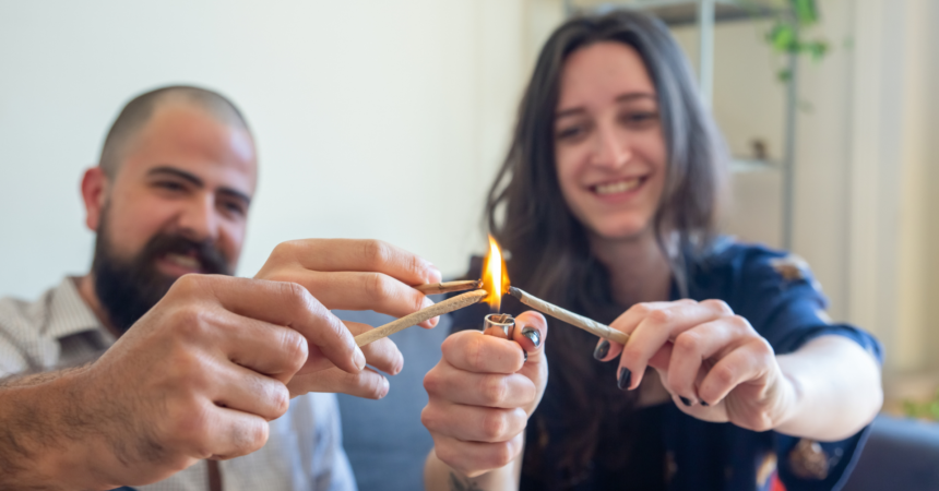 Three friends light joints together over a lighter flame. One is a woman with brunette hair, smiling